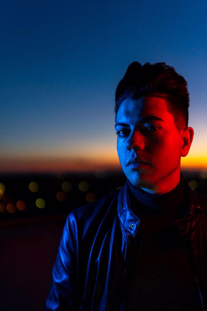 Serious young male with stylish haircut wearing black leather jacket looking away while standing on street against dark sunset sky