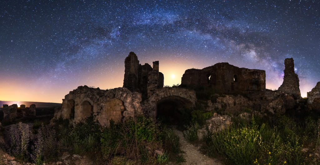 Aged ruined building among green field plants under colorful starry night sky with milky way