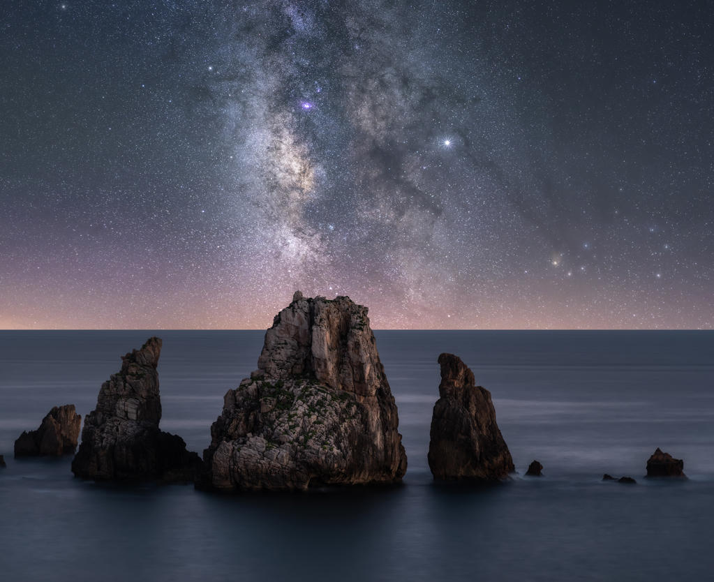 Big rough cliffs on blue calm ocean during bright evening under colorful starry sky with milky way