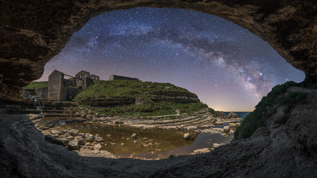 Stone rough cave on seashore with stone aged ruins and green hills under night sky with milky way and stars