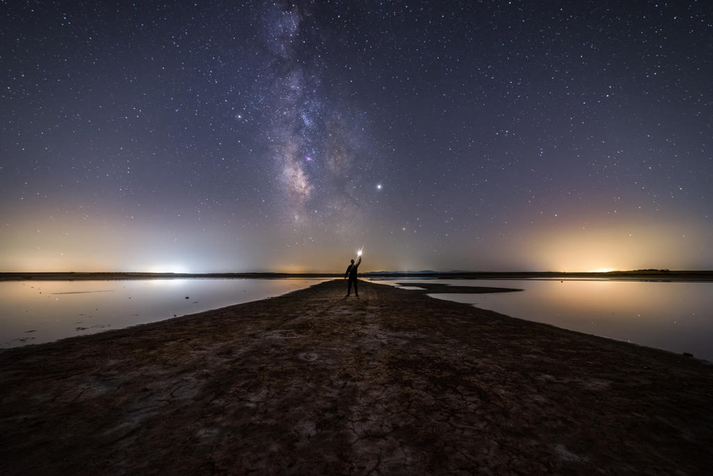 Back view of anonymous man standing on empty road among calm water and reaching out to star under colorful nigh sky with milky way on background
