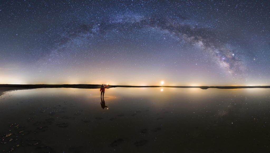 Silhouette of anonymous man standing on reflection surface of water and reaching out to starry colorful night sky with milky way