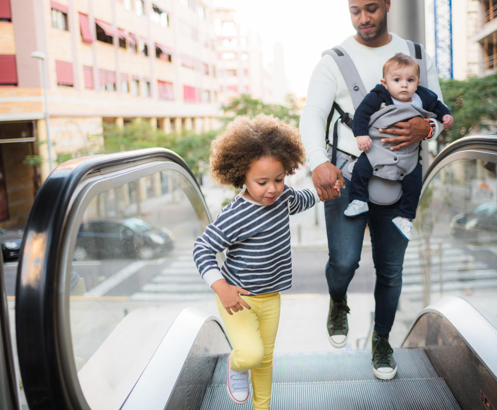 From above of positive young ethnic man carrying infant son and holding hand of cheerful little daughter while standing on escalator in city