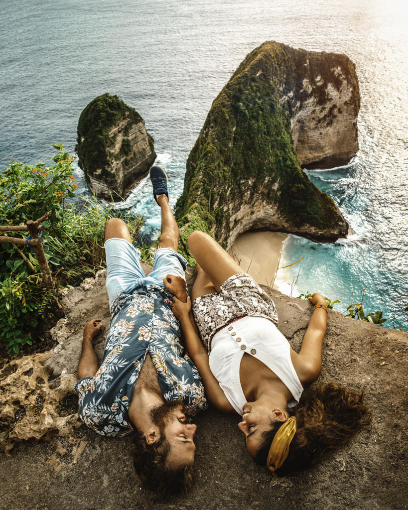 Carefree young couple on vacation in light casual wear lying down on peak of rough cliff and looking at each other with breathtaking landscape on background in Indonesia