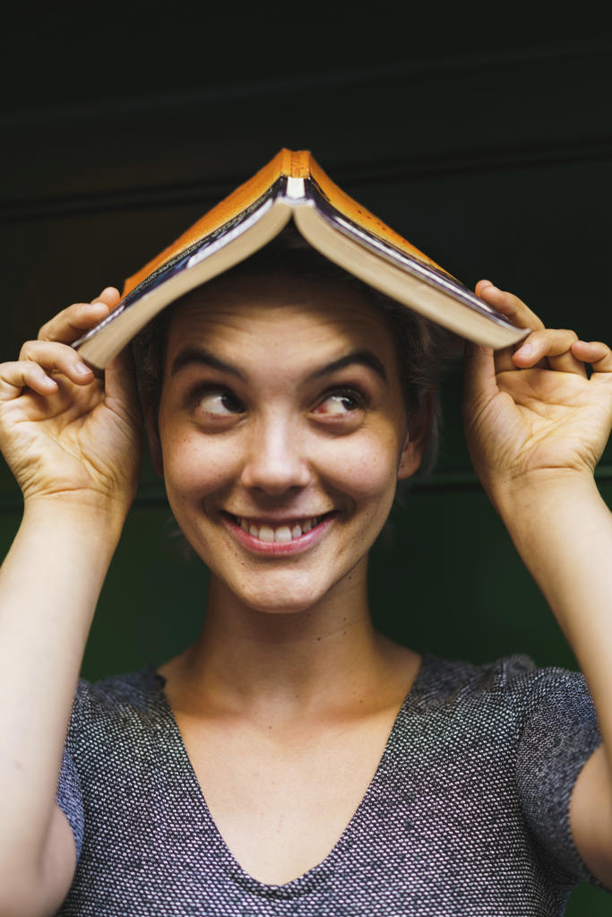 Portrait of young playful woman smiling away and holding opened book on head.