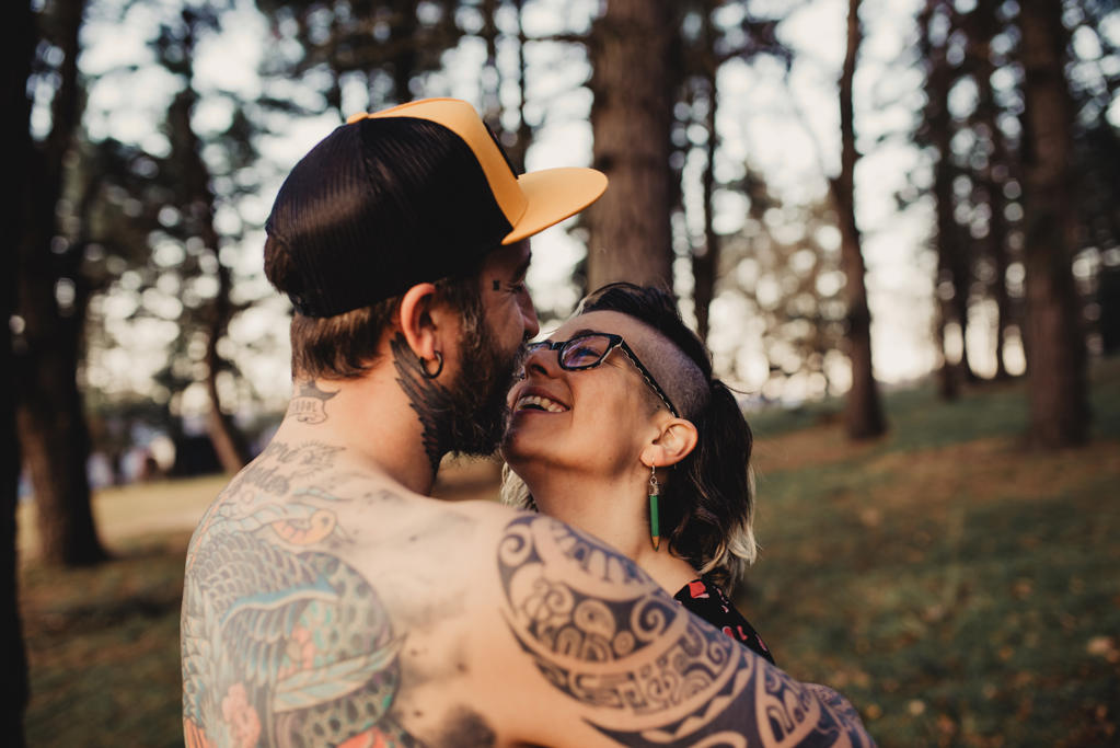 Back view of young shirtless man in tattoos with snapback embracing woman in park on blurred background