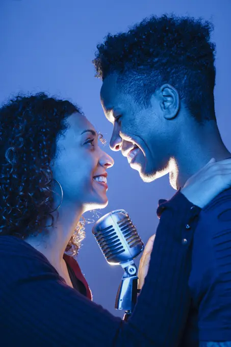Side view of young African man and attractive woman holding vintage microphone and embracing in blue light