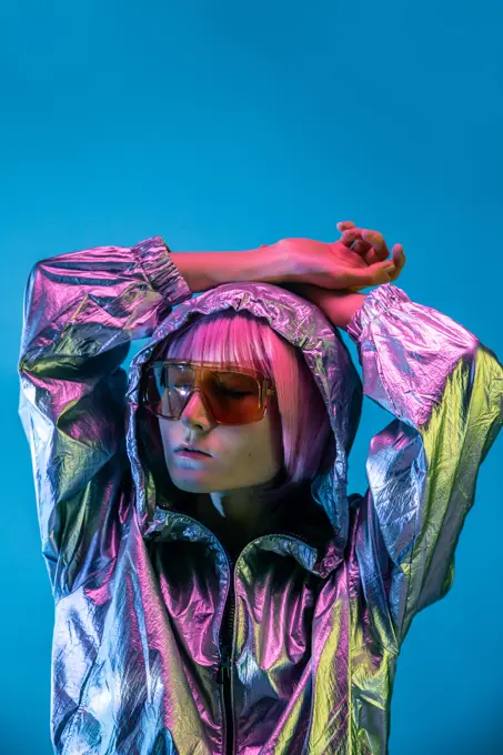Trendy young Japanese woman with purple hair standing in sparkly silver jacket and red sunglasses on blue background