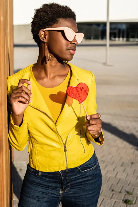 Trendy African American woman in sunglasses in yellow jacket enjoying heart shaped lollipop by wooden fence 