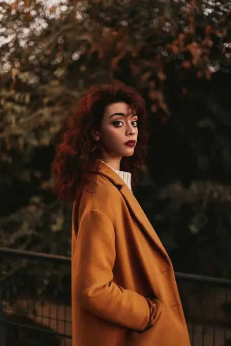 Side view of young sensual female with dark curly hair wearing knitted jumper and overcoat standing in park putting hand on metal railing while looking at camera with thoughtful look