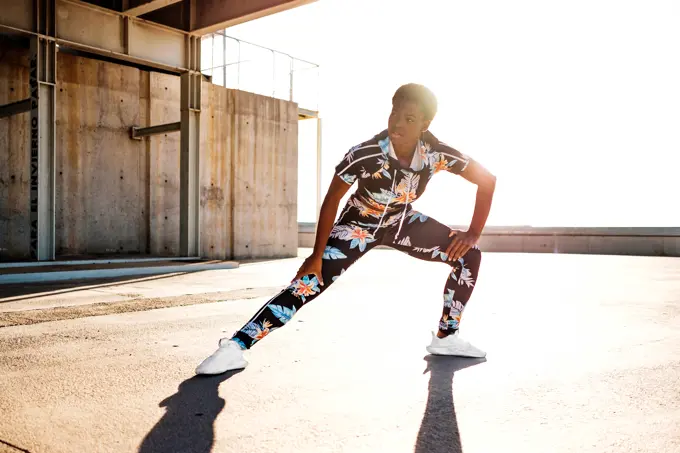 Focused African American adult woman in flowered sportswear stretching leg muscles while standing alone and warming up before training among urban environment in sunny day