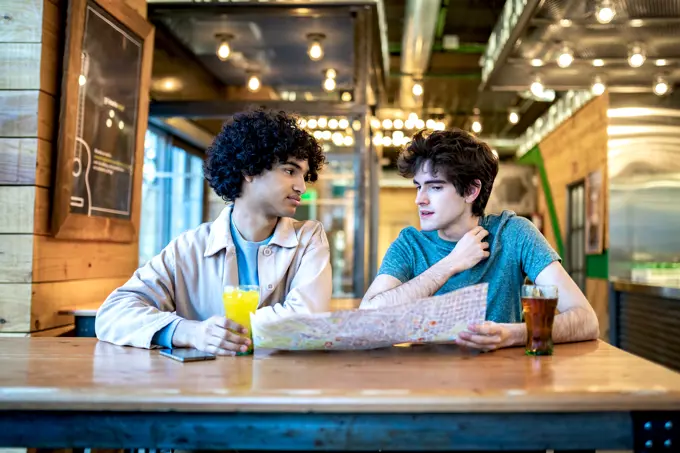 Multiethnic young homosexual men with direction navigation map and fresh drinks smiling looking at each other while sitting at cafe table during romantic date