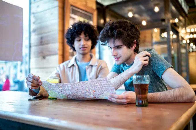 Multiethnic young homosexual men looking at direction navigation map and fresh drinks smiling while sitting at cafe table during romantic date