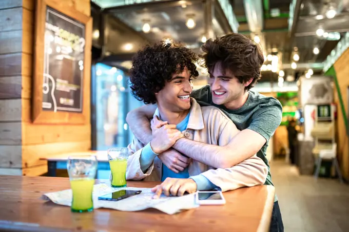 Multiethnic young homosexual men embracing each other looking at direction navigation map and having fresh drinks smiling while sitting at cafe table during romantic date