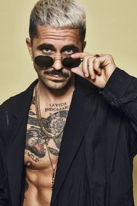 Portrait of brutal muscular sexy fit male with tattooed torso wearing black coat and stylish sunglasses and accessories standing against beige background looking at camera
