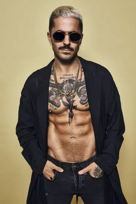Brutal muscular sexy fit male with tattooed torso wearing black coat and trendy ripped jeans with stylish sunglasses and accessories standing against beige background looking at camera