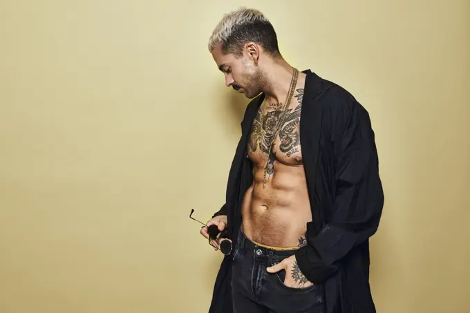 Brutal muscular sexy fit male with tattooed torso wearing black coat and trendy jeans with stylish sunglasses and accessories standing against beige background looking away