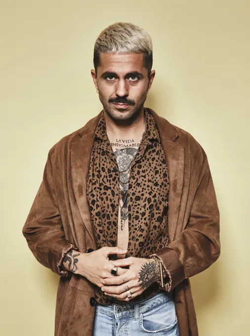 Fashionable male model with tattoos wearing trendy coat over leopard shirt and jeans standing against beige background and looking at camera