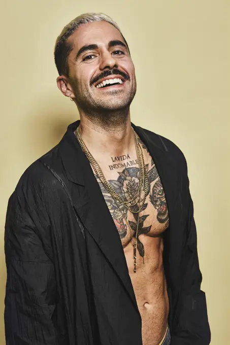 Confident cheerful stylish man with mustache showing off his muscular tattooed torso wearing black coat looking at camera against beige background