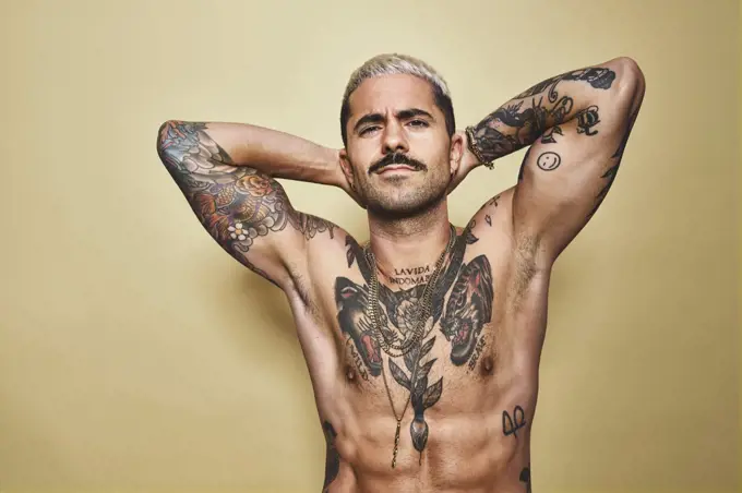 Handsome sexy muscular male with various tattoos on naked torso and arms looking at camera while standing against beige background