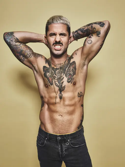 Handsome sexy attractive muscular male with various tattoos on naked torso and arms looking at camera making faces with mouth while standing against beige background