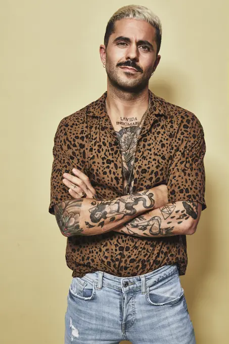Fashionable male model with tattoos wearing trendy leopard shirt and jeans standing against beige background and looking at camera with arms crossed