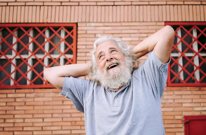 Aged bearded male in cotton tee shirt smiling and looking away while standing with hands behind head near loft building