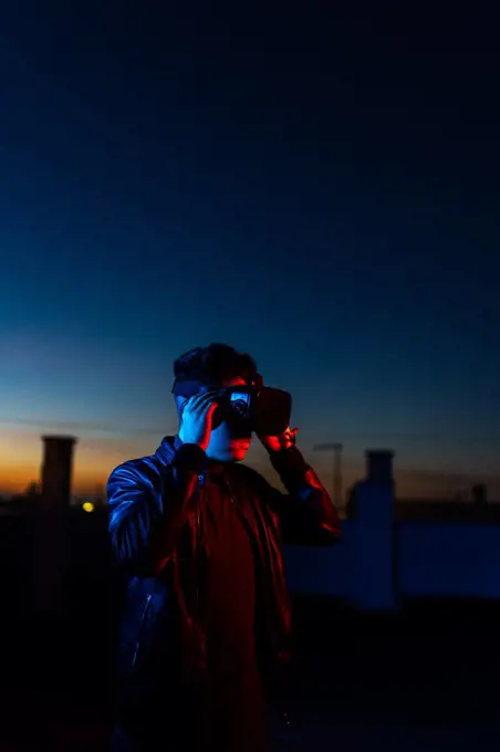 Modern male with VR headset exploring virtual world while standing in red neon light on street against dark sunset sky