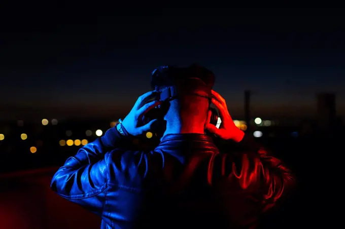 Back view of anonymous male in black leather jacket and virtual reality goggles standing on dark street against night sky with city lights in background