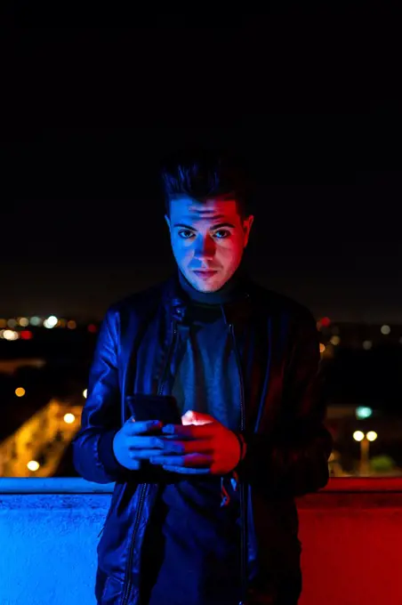 Young male browsing at smartphone looking at camera while standing on balcony under red and blue light against illuminated city street and dark sky at night