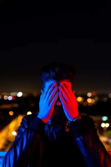 Contemporary young guy covering face with hand while standing under bright red and blue light on blurred background of city street at night