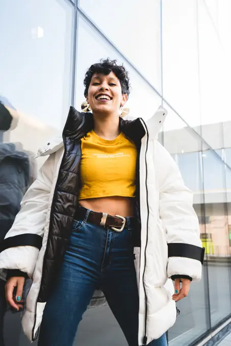 From below of joyful ethnic female teenager in colorful casual clothing and warm jacket laughing and looking at camera with modern building on background