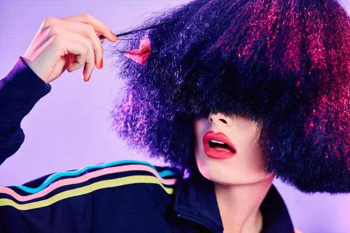 Astonished female in stylish sportswear inspecting fake hair under bright illumination against violet background