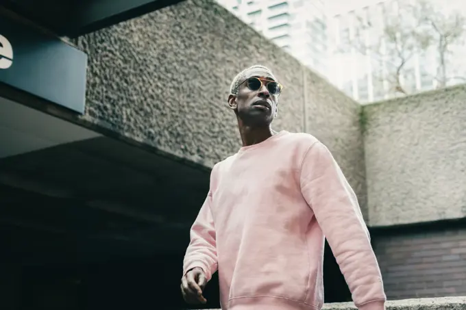 Low angle of confident black guy in stylish pink sweatshirt and sunglasses looking away and walking near underground entrance on city street