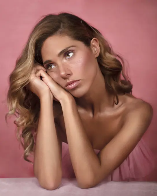 Pretty young woman clasping hands under cheek and looking away while sitting against pink background and dreaming