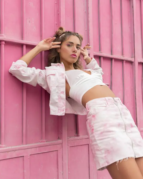 Slim stylish female in casual clothes pointing at temples with hand gun gesture and looking at camera while leaning on pink wall on city street