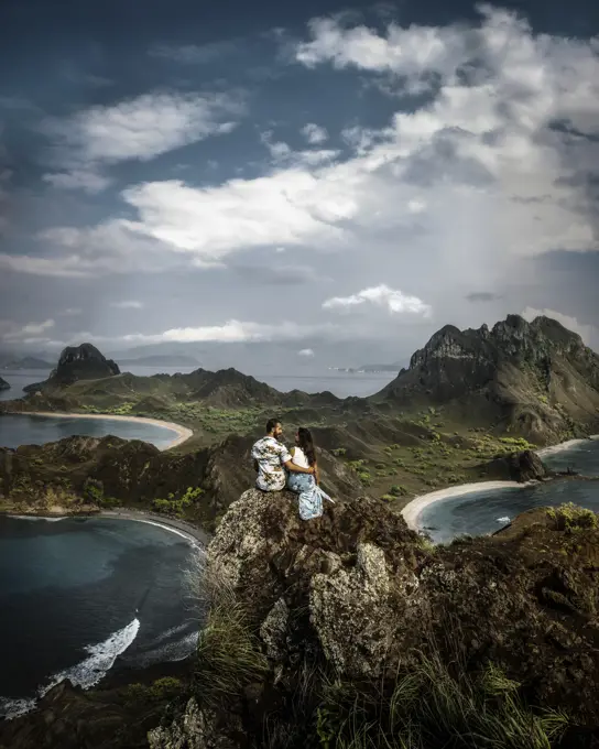 Side view of carefree young couple on vacation in light casual wear standing on peak of rough cliff and looking at each other with breathtaking landscape on background in Indonesia