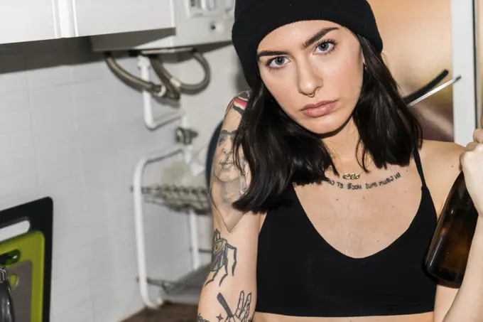 Young tattooed brunette in hat and black bra holding glass bottle and looking at camera on kitchen.
