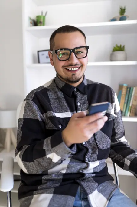 Cheerful man in glasses sitting in office and using smartphone.