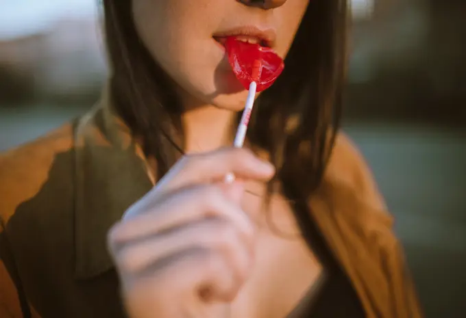 Crop young woman eating red colored heart-shaped sweet lollipop.