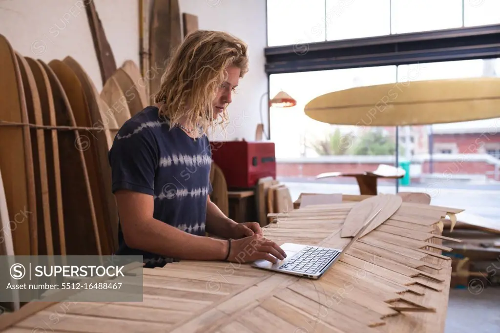 Caucasian male surfboard maker in his studio, working on a project using his laptop, with surfboards in a rack in the background. 