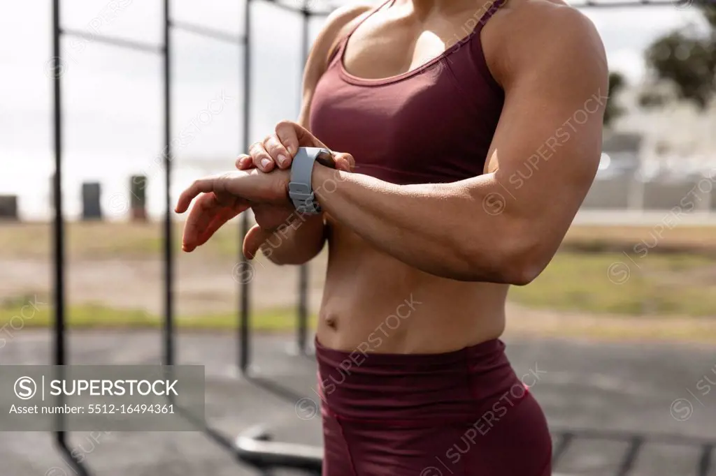 Front view mid section of a sporty Caucasian woman exercising in an outdoor gym during daytime, looking at her smartwatch.
