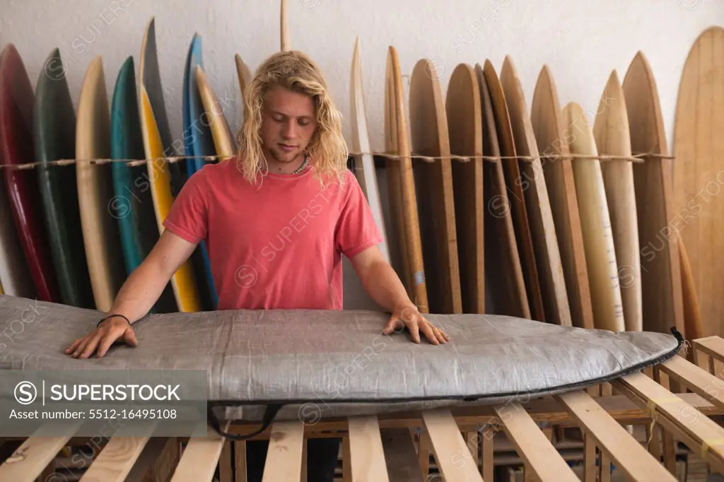 Caucasian male surfboard maker working in his studio, inspecting a surfboard covered with a grey case, with surfboards in a rack in the background.