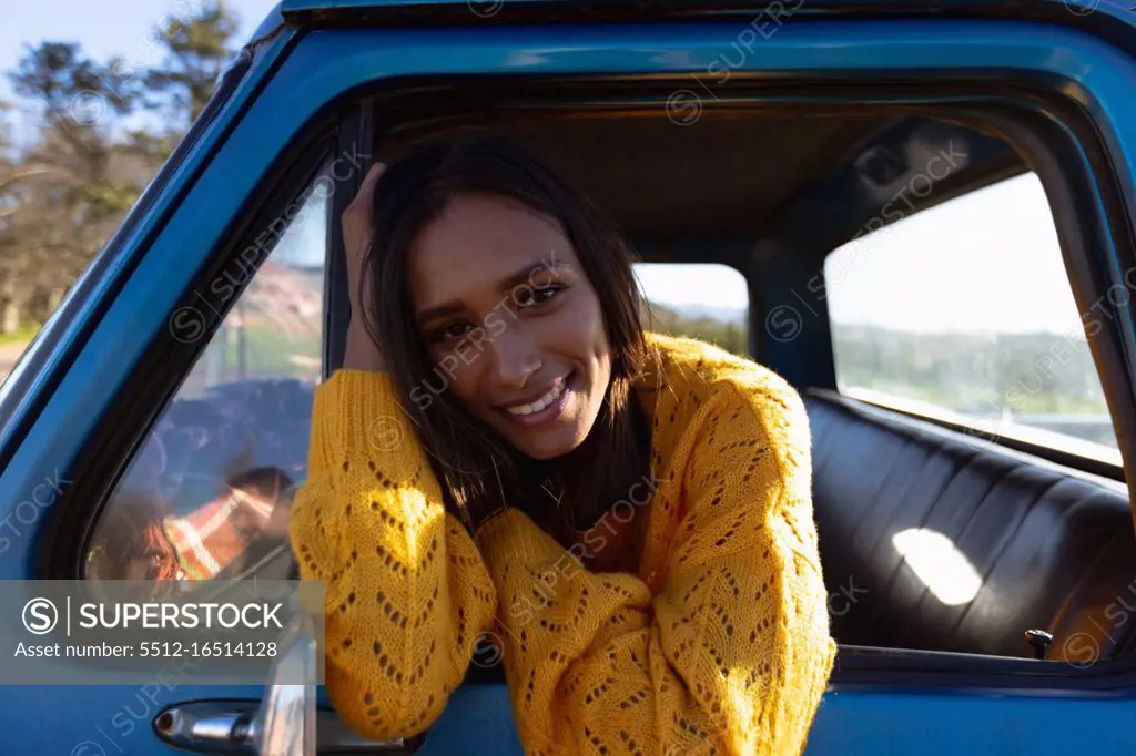 Portrait close up of a young mixed race woman sitting in the front passenger seat of a pick-up truck, leaning out of the side window smiling to camera during a road trip