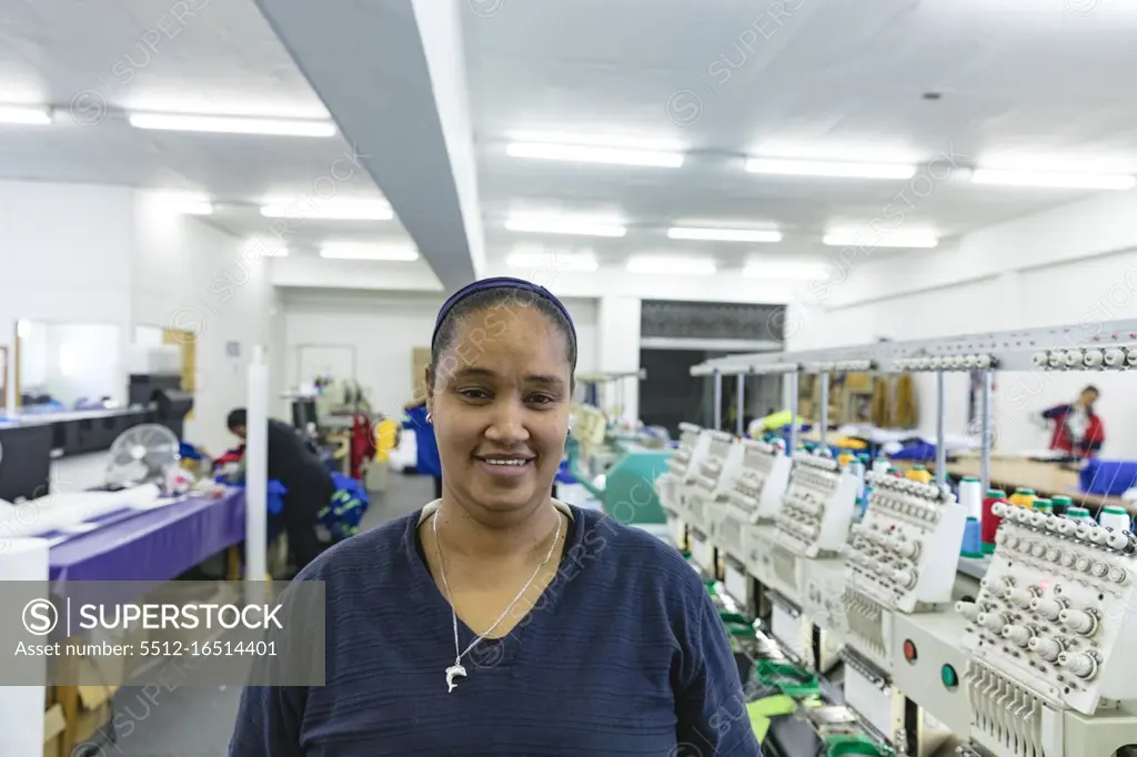 Portrait close up of a young mixed race woman standing beside rows of machines in a brightly lit sports clothing factory, looking to camera and smiling. In the background some of coworkers can be seen. They are working in a clothing factory.