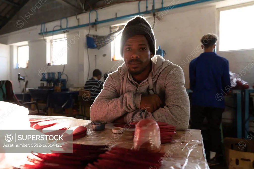 Portrait close up of a young African American man sitting at a workbench in a factory makimg cricket balls, looking to camera and smiling. In the background colleagues are working on other parts of the production line. They are working in a clothing factory.