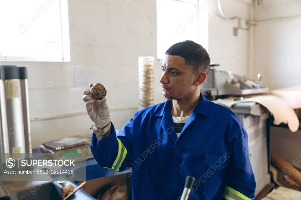 Front view close up of a young mixed race man wearing gloves and overalls holding the core of a ball and checking it at a factory making cricket balls, with equipment and materials visible in the background. They are working in a clothing factory.