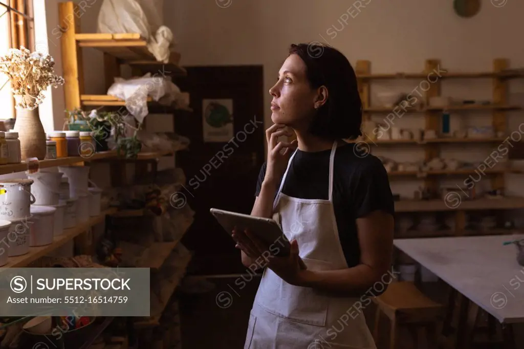 Side view a young Caucasian female potter with her hand on her chin looking out of the window and using a tablet in a pottery studio