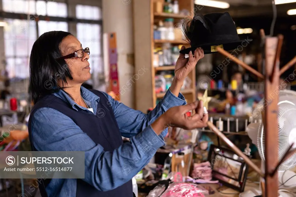 Side view close up of a middle aged mixed race woman inspecting and adding the finishing touches to a hat on a stand in the workshop at a hat factory, with materials visible in the background