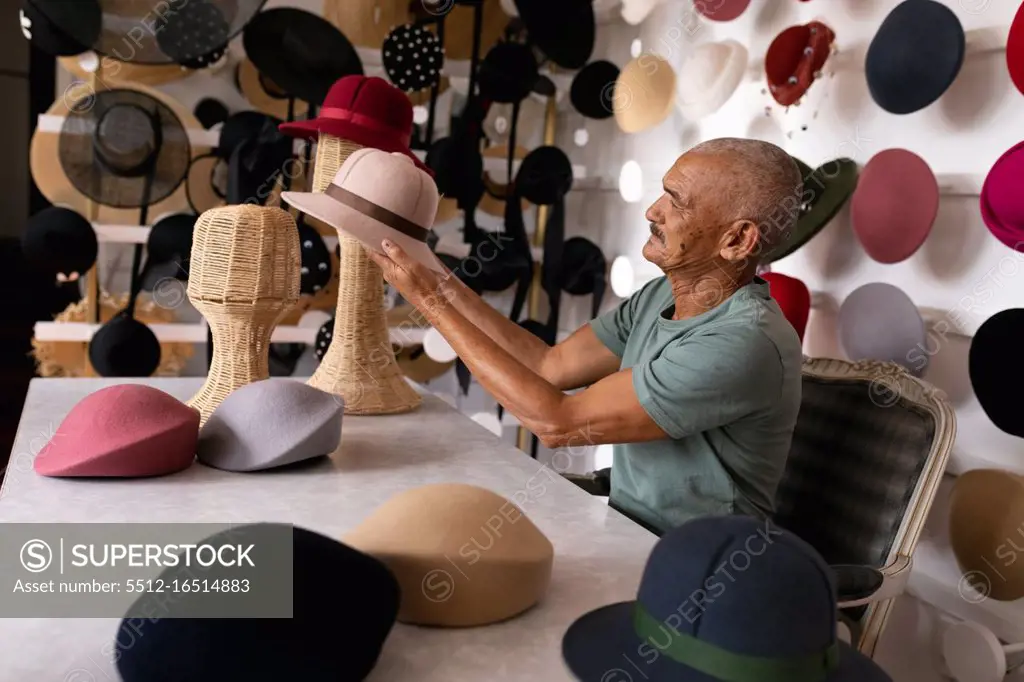 Side view of a senior mixed race man sitting and inspecting a finished hat, surrounded by hats on display in the showroom at a hat factory